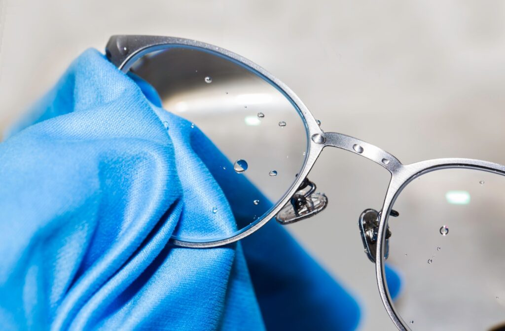 A close-up image of a pair of gray eyeglasses with drops on the lens and a blue microfiber cloth cleaning them.