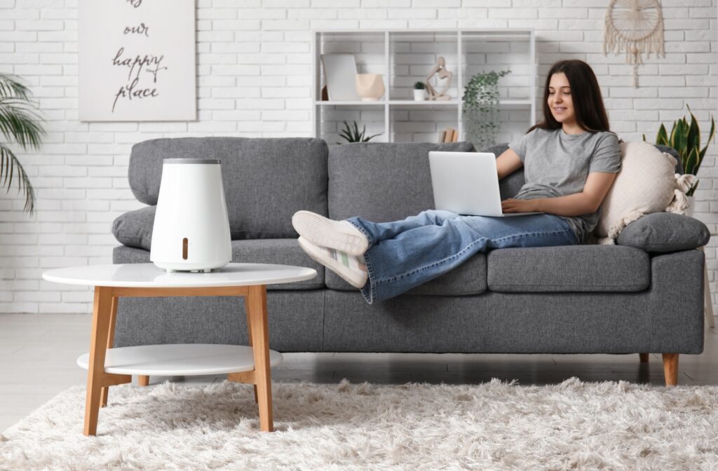 A person sits on a couch with a laptop while a humidifier runs on the table nearby.
