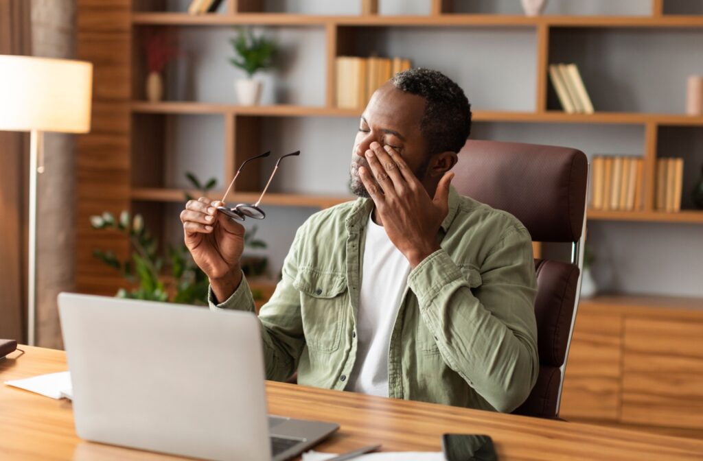 A person rubs their eyes while working at a desk with a laptop and glasses in hand.