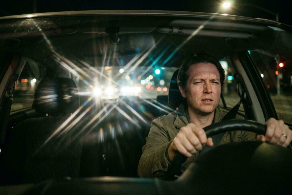 An adult driving a car at night squints as bright lights shine through the windshield.